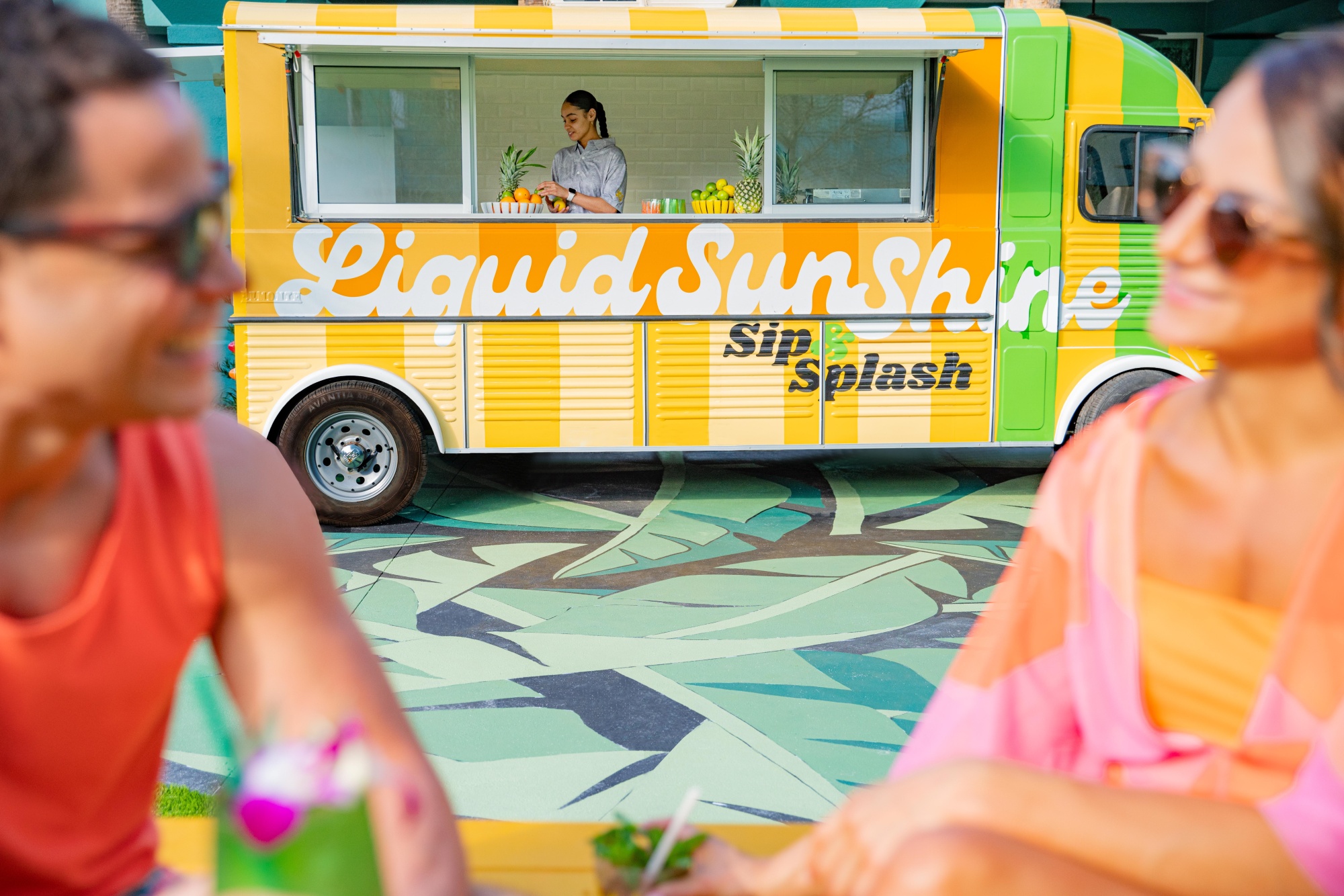 A bright and colorful mobile food truck with 'Liquid Sunshine' painted on the side. In the foreground a couple is enjoying cocktails.